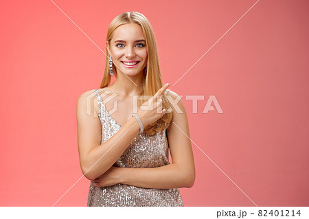 Confident attractive feminine glamour blond young woman in silver glittering dress smiling broadly pointing up discuss recent news promo sales standing friendly grinning camera red background Confident attractive feminine glamour blond young woman in silver glittering dress smiling broadly pointing up discuss recent news promo sales standing friendly grinning camera red background 82401214