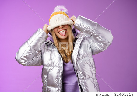 Playful carefree funny blond girl having fun fool around careless what people thing acting immature joyfully pulling hat eyes sticking tongue smiling standing silver jacket purple background Playful carefree funny blond girl having fun fool around careless what people thing acting immature joyfully pulling hat eyes sticking tongue smiling standing silver jacket purple background 82401215