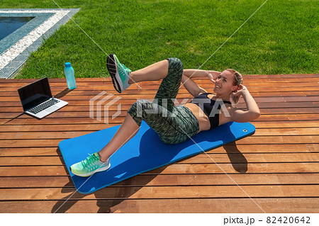A beautiful European girl, performs exercises for abdominal, remote lessons, a fitness trainer on a laptop. In the park With pool.on the grass. A beautiful European girl, performs exercises for abdominal, remote lessons, a fitness trainer on a laptop. In the park With pool.on the grass. 82420642