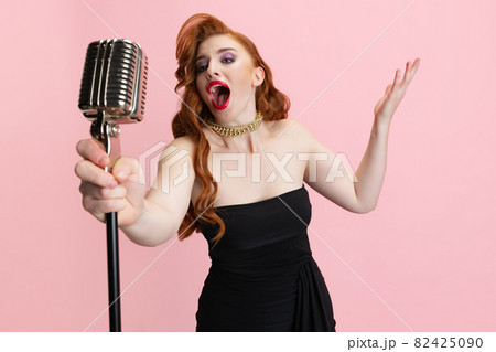 Cropped studio portrait of expressive beautiful woman singing in microphone isolated over pink background. Retro music performer Cropped studio portrait of expressive beautiful woman singing in microphone isolated over pink background. Retro music performer 82425090
