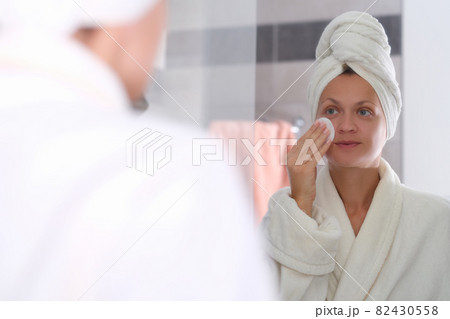 Young woman in bathrobe and towel on head washing off cosmetics from face in front of mirror 82430558