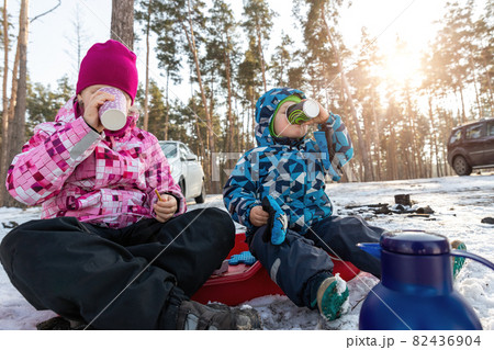 Two cute adorable beautiful caucasian little siblings children boy girl enjoy drinking warm drink tea or chocolate in paper cup and eat on bright sunny winter morning forest family picnic outside Two cute adorable beautiful caucasian little siblings children boy girl enjoy drinking warm drink tea or chocolate in paper cup and eat on bright sunny winter morning forest family picnic outside 82436904