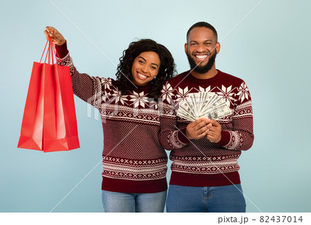 Excited african american spouses in Christmas sweaters holding shopping bags and bunch of money, blue background 82437014