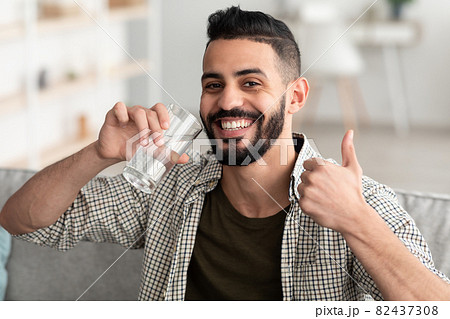 Positive Arab guy drinking fresh clear water from glass, showing thumb up gesture and smiling at camera at home 82437308