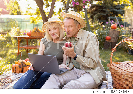 Happy senior spouses web surfing on internet with laptop, sitting on blanket in garden, enjoying sunny autumn day Happy senior spouses web surfing on internet with laptop, sitting on blanket in garden, enjoying sunny autumn day 82437643
