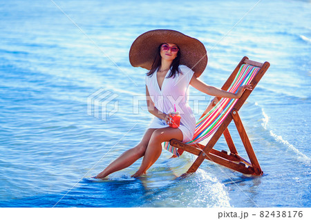 girl in sunhat and tunic sitting on the beach chair at seaside while holds cocktail. Summer vacation 82438176