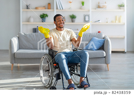 Happy disabled black man in wheelchair holding detergent and rag, ready to clean his home 82438359