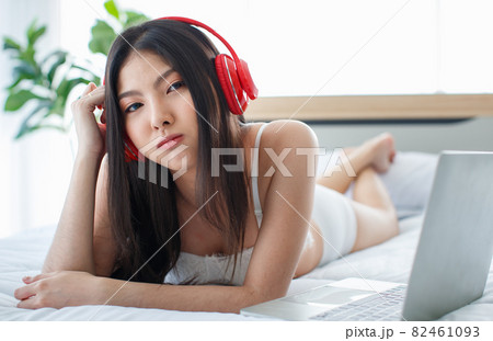 Beautiful young naughty sexy girl with long hair and white romantic underwear lying down at bed end beside laptop wearing red earphone for music listening and glacing at someone Beautiful young naughty sexy girl with long hair and white romantic underwear lying down at bed end beside laptop wearing red earphone for music listening and glacing at someone 82461093