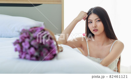 Beautiful young sexy girl with romantic pajamas sit beside luxury white bed elegantly decorated by beautiful bunch of purple flower and glance seductively to someone at bed end 82461121