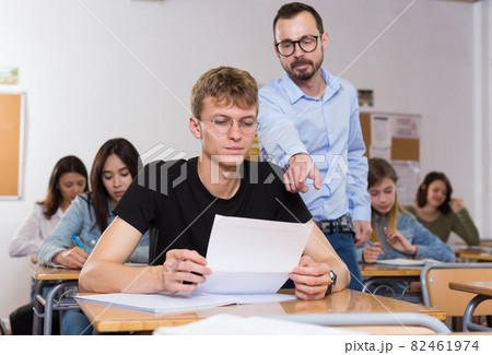 Schoolboy is writing answer the question on paper and teacher explaining it Schoolboy is writing answer the question on paper and teacher explaining it 82461974