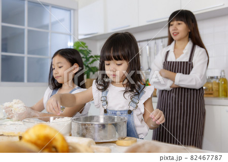 Two little cute Asian girls, 3 years and 7 years old, learning how to make bread and bakery with a curious and happy smiley face. Their mom standing and looking behind them Two little cute Asian girls, 3 years and 7 years old, learning how to make bread and bakery with a curious and happy smiley face. Their mom standing and looking behind them 82467787
