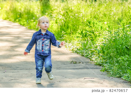 Blond-haired girl baby runs along the asphalt path in the park on a sunny day. 82470657