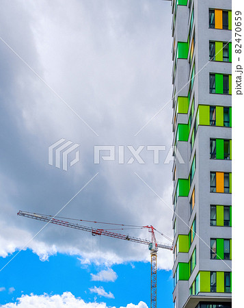 A tall construction crane next to a modern building against a blue sky and cloud 82470659