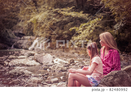 Happy mother with a child sitting on the grass on a background of forested mountains Happy mother with a child sitting on the grass on a background of forested mountains 82480992