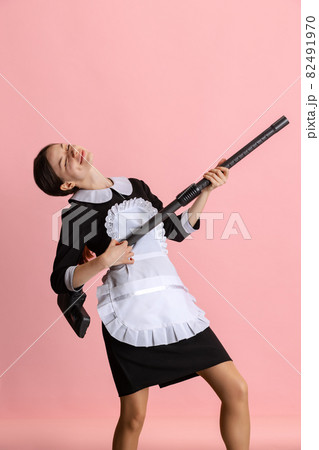 Dreamy girl, chambermaid pretending to be a musician, holding vaccum cleaner like guitar isolated over pink background 82491970