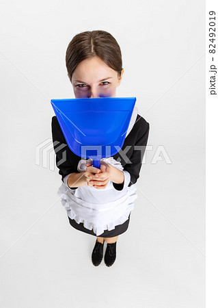 Pretty young girl in cleaning uniform hiding face behind dustpan isolated over white background 82492019