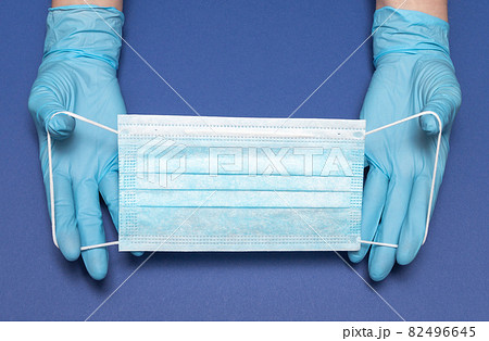 Hands in blue medical gloves is holding face mask on blue background. Protective respiratory medical mask against spreading virus. Close up, copy space, top view, flat lay. Hands in blue medical gloves is holding face mask on blue background. Protective respiratory medical mask against spreading virus. Close up, copy space, top view, flat lay. 82496645