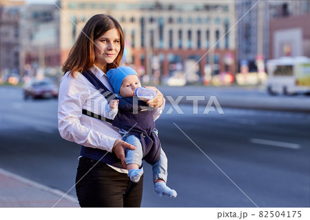 Mother is feeding baby in sling with bottle on street. 82504175
