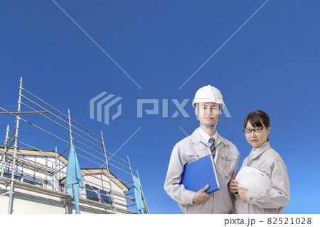 青空と一戸建て工事現場を背景に立つ、作業着を着たカメラ目線の男女技師 82521028
