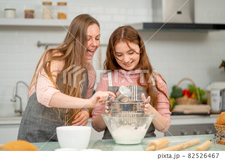 Caucasian mother with apron cheer her daughter to process of cake cooking by sift flour and enjoy Caucasian mother with apron cheer her daughter to process of cake cooking by sift flour and enjoy 82526467