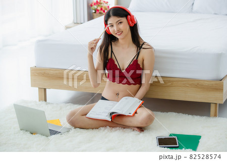 Beautiful young Asian sexy girl on red bra sit back against white bed end happily smile as she enjoy reading book and studying lecture media from laptop, tablet, and red earphone in bright morning 82528574
