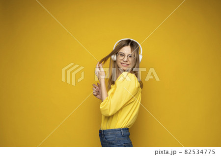 Beautiful attractive young blond woman wearing yellow t-shirt and glasses in white headphones listening music, dancing and laughing on blue background in studio. Relaxing and enjoying. Lifestyle. 82534775