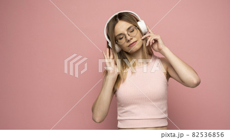 Beautiful attractive young blond woman wearing pink t-shirt and glasses in white headphones listening music and smiling on pink background in studio. Relaxing and enjoying. Lifestyle. 82536856