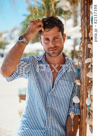 Adult man in striped shirt touching hair and looking at camera while leaning on wooden pole with seashells on sunny summer day on beach 82537398