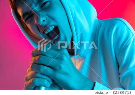 Portrait of young man, student or boy in casual clothes singing with microphone isolated on magenta studio backgroud in neon. 82539713
