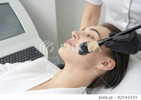 Cosmetologist applying black mask on the face of a beautiful mature woman for carbon peeling 82543335