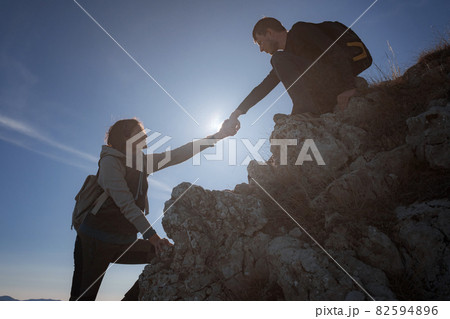 Silhouettes of two people climbing mountains and helping against the blue sky. 82594896