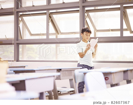 asian elementary schoolchild standing by the windows reading a book in classroom asian elementary schoolchild standing by the windows reading a book in classroom 82607367