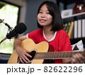 Happy cheerful pretty smiling of portrait a Asian girl vocalist with a Guitar recording a song front of microphone in living room. 82622296