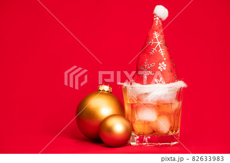 Glass of whiskey with red santa hat and christmas baubles on red background christmas drink concept Glass of whiskey with red santa hat and christmas baubles on red background christmas drink concept 82633983