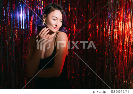 Merry christmas. Festive woman. Holiday look. Model shooting. Pretty smiling lady purple curly hair posing on shimmering red cascade curtain background blur shadow light copy space. Merry christmas. Festive woman. Holiday look. Model shooting. Pretty smiling lady purple curly hair posing on shimmering red cascade curtain background blur shadow light copy space. 82634175