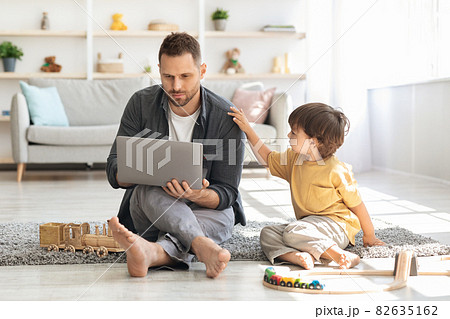Young concentrated man working on laptop at home, sad little boy touching his shoulder, calling daddy to play with him Young concentrated man working on laptop at home, sad little boy touching his shoulder, calling daddy to play with him 82635162