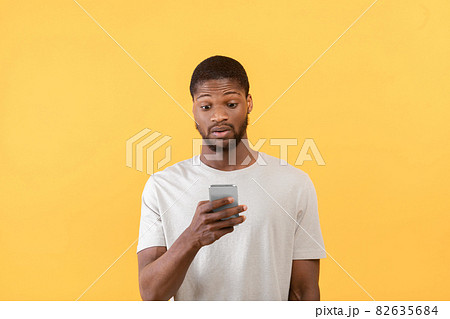 Emotional african american guy looking at smartphone, reading shocked message of promo or offer, yellow background Emotional african american guy looking at smartphone, reading shocked message of promo or offer, yellow background 82635684