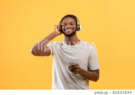 Excited young african american man listening music and holding smartphone, standing over yellow background Excited young african american man listening music and holding smartphone, standing over yellow background 82635834