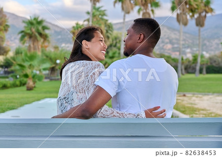 Beautiful young multi-ethnic couple sitting together on a bench in a tropical park Beautiful young multi-ethnic couple sitting together on a bench in a tropical park 82638453