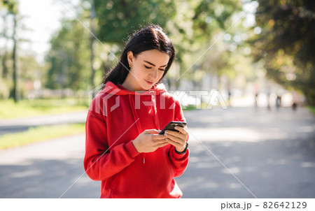 Stylish girl in red sweater posing on alley holding smartphone. Happy woman with beautiful smile chatting on cell smartphone while relaxing in the park, outdoors Stylish girl in red sweater posing on alley holding smartphone. Happy woman with beautiful smile chatting on cell smartphone while relaxing in the park, outdoors 82642129