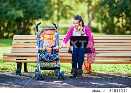 Woman with child in baby carriage makes remote work in park. 82651515