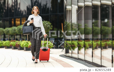 Young happy stylish business woman in formal outfit with suitcase walking along city street Young happy stylish business woman in formal outfit with suitcase walking along city street 82655289
