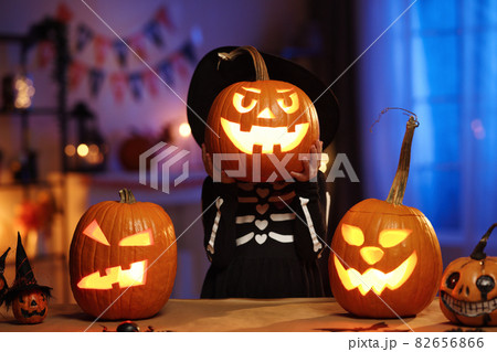 Little girl in Halloween costume holding glowing jack-o-lantern carved pumpkin in front of face 82656866