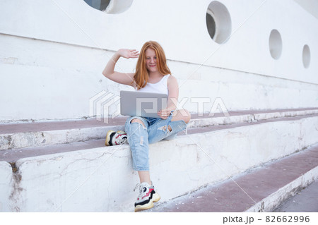 Red-haired teen girl talking on video call while sitting on steps with laptop Red-haired teen girl talking on video call while sitting on steps with laptop 82662996