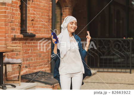 Middle eastern woman in hijab listening music with headphones and dancing outdoors. Woman independence and feminism concept. Copy space. Middle eastern woman in hijab listening music with headphones and dancing outdoors. Woman independence and feminism concept. Copy space. 82663922