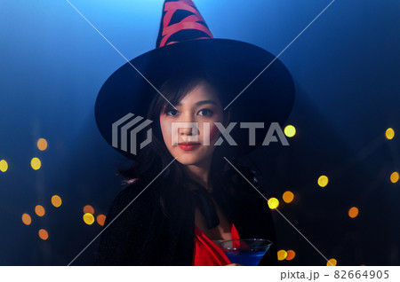 Portrait of beautiful young asian woman in witch halloween costumes drinking cocktail in halloween party over dark magic background - holiday party concept. Portrait of beautiful young asian woman in witch halloween costumes drinking cocktail in halloween party over dark magic background - holiday party concept. 82664905