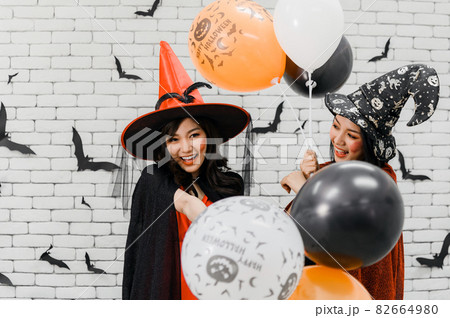 Two beautiful young asian woman in witch halloween costume holding balloon on white brick background decorated with black paper bat shape for halloween party Two beautiful young asian woman in witch halloween costume holding balloon on white brick background decorated with black paper bat shape for halloween party 82664980