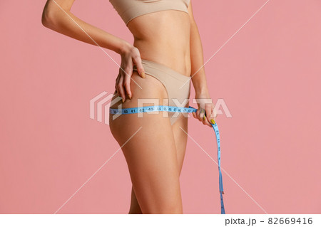 Close-up slim female body with well kept skin in pastel color panties using centimeter isolated over pink studio background. 82669416
