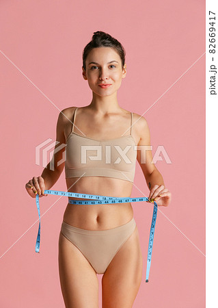 Portrait of young beautiful slim girl in lingerie takes measurements from yourself isolated over pink studio background. 82669417