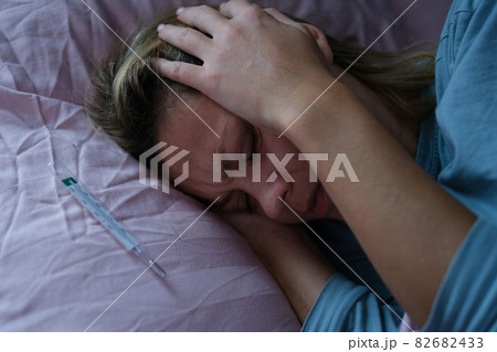 Woman lying in bed and holding sore head with hands near mercury thermometer 82682433